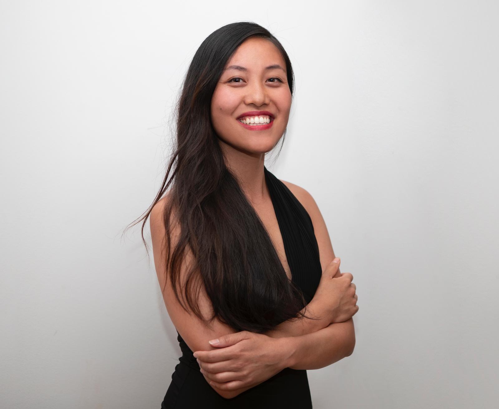 Portrait of Emily Sha on a light grey background, she is standing smiling with her arms crossed, she is wearing a black v neck dress